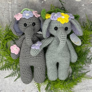 May include: Two gray crocheted elephant plushies with flower crowns. One elephant has a pink and purple flower crown, the other has a yellow and purple flower crown.
