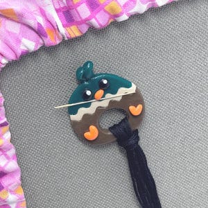 May include: A handmade polymer clay pin in the shape of a teal and brown donut with orange heart accents. A silver pin is attached to the back. A black tassel hangs from the bottom. The pin is on a gray surface.