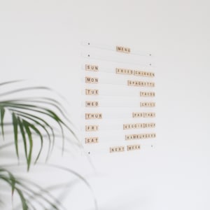 Puede incluir: Una pared blanca con un menú semanal escrito en ella usando fichas de letras. El menú incluye pollo frito, espaguetis, tacos, chili, pad thai, sopa de verduras, hamburguesas y la próxima semana.