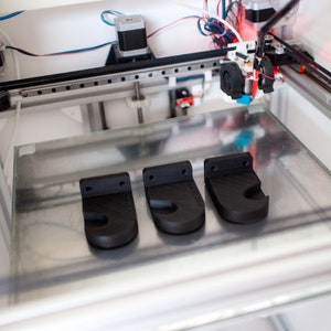 May include: Three black plastic wall hooks printed on a 3D printer. The hooks are on a glass surface inside the printer.