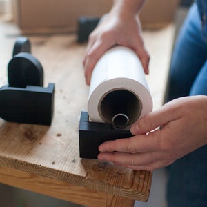 May include: A person is holding a roll of white paper, which is being placed into a black plastic holder. The holder is on a wooden table.