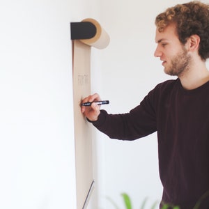 May include: A person writing on a brown paper roll mounted on a wall. The paper roll is attached to a black metal holder. The person is writing the words "From the" on the paper roll.