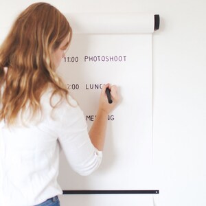 May include: A person writing on a white roll-up wall calendar with a black marker. The calendar has the following text: 11:00 PHOTOSHOOT, 2:00 LUNCH, MEETING.