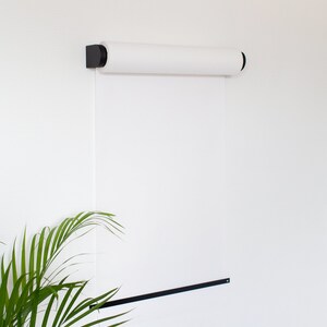 May include: A white roll of paper mounted on a white wall with black brackets. A black metal bar is attached to the wall below the paper roll. A green plant is in the lower left corner of the image.