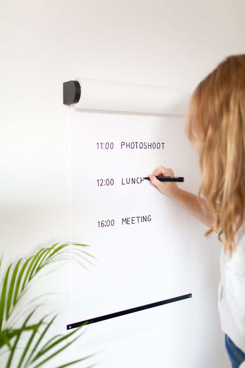 May include: A white roll-up whiteboard with a black marker and a handwritten schedule. The schedule includes the times 11:00, 12:00, and 16:00 with the events "PHOTOSHOOT", "LUNCH", and "MEETING".