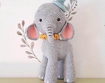 May include: A handmade gray crochet elephant toy with a light blue hat and a yellow collar. The elephant has pink inner ears and is standing on a green surface. The background features a white wall with floral decals.