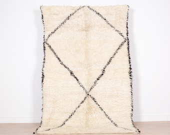 May include: Cream-colored, rectangular rug with a large black diamond pattern. The rug has a shaggy texture and a fringe along the edges. The design is simple yet striking, suitable for various interior styles.