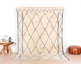 May include: Cream-colored, shaggy rug with a geometric diamond pattern in black. The rug is displayed against a white wall, showcasing its texture and design. A wooden side table and leather ottoman are in the background.
