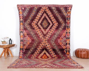 May include: A large, rectangular rug with a geometric diamond pattern in shades of burgundy, orange, and pink. The rug has a textured appearance and a fringe along the bottom edge. The rug is displayed on a light-colored floor.