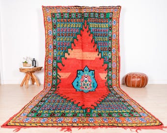Könnte beinhalten: Ein großer, leuchtend roter Teppich mit einem zentralen geometrischen Design in Türkis, Blau und Gelb. Der Teppich hat einen detaillierten Rand mit Blumenmustern und Fransen an den Kanten. Der Teppich wird auf einem hellen Holzboden präsentiert.
