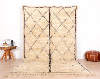 May include: A large, cream-colored rug with a geometric diamond pattern in dark brown. The rug has a thick pile and fringe along the bottom edge. The rug is displayed on a light-colored floor.