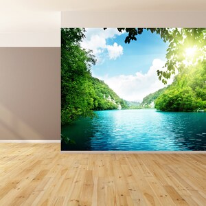 May include: A scenic landscape photograph featuring a lake surrounded by lush green trees and hills. The water is a vibrant blue, and the sky is a clear blue with white clouds. The sun is shining brightly, creating a warm and inviting atmosphere.