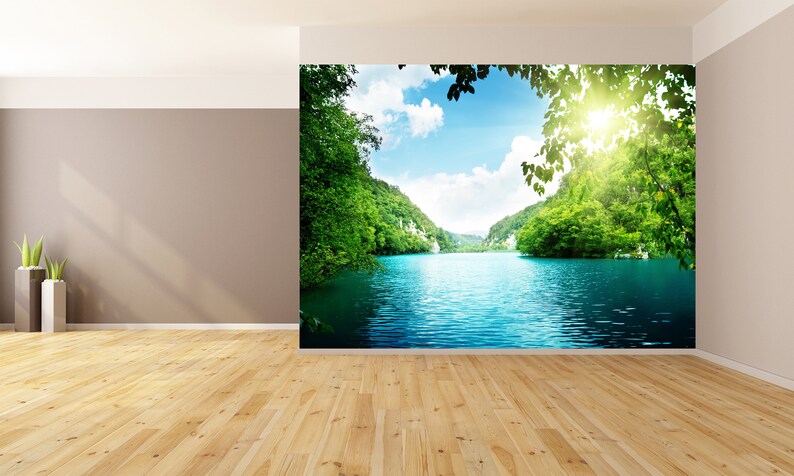 May include: A scenic landscape photograph featuring a lake surrounded by lush green trees and hills. The water is a vibrant blue, and the sky is a clear blue with white clouds. The sun is shining brightly, creating a warm and inviting atmosphere.