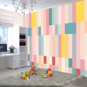 Op de afbeelding: Een speelkamer voor kinderen met een kleurrijke geometrische muurschildering. De muur is geschilderd in tinten roze, geel, blauw en groen. Er ligt een teddybeer, een speelgoed en houten blokken op de vloer voor de muur.