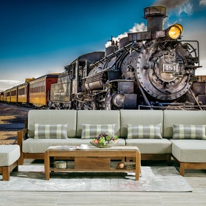 May include: A vintage steam locomotive train with red and yellow passenger cars against a blue sky. The train is in front of a gray couch with plaid pillows and a wooden coffee table.