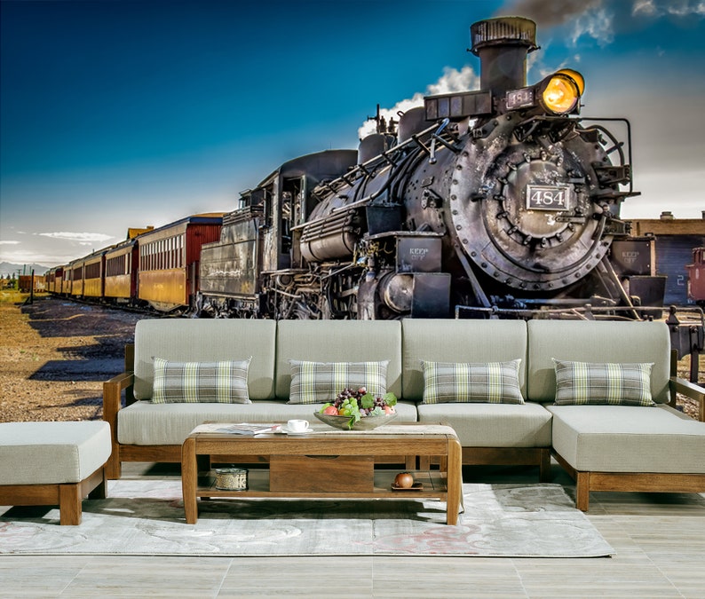 May include: A vintage steam locomotive train with red and yellow passenger cars against a blue sky. The train is in front of a gray couch with plaid pillows and a wooden coffee table.