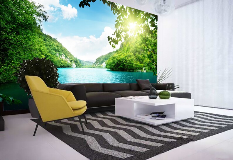 May include: A living room interior design featuring a large wall mural depicting a scenic lake surrounded by lush green trees and a bright blue sky. The room includes a yellow armchair, a black sofa, a white coffee table, and a black and white patterned rug.