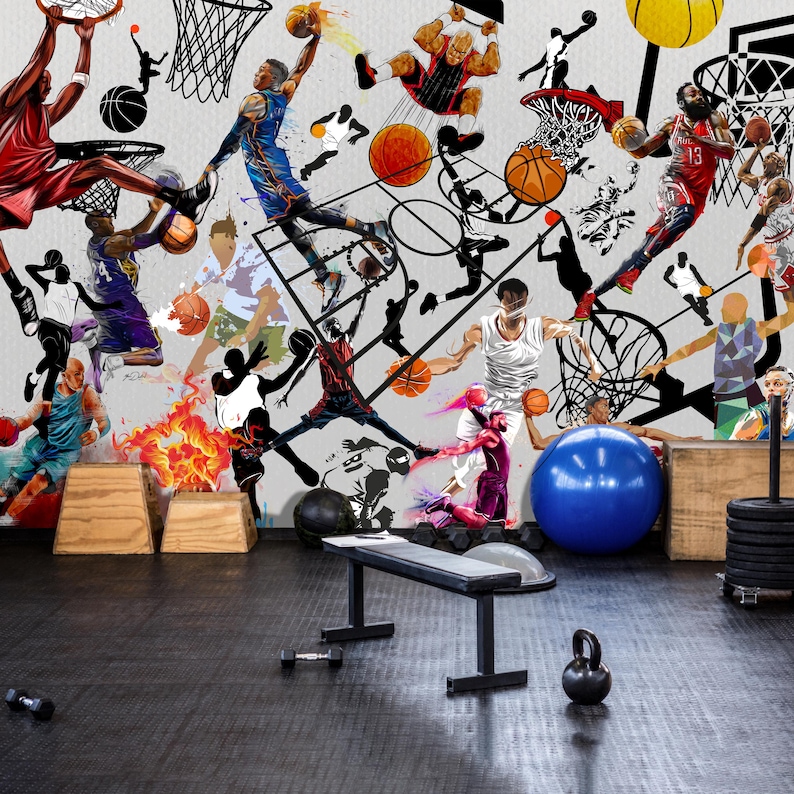 A gym interior with a wall mural featuring a basketball theme. The mural is a collage of basketball players in various poses, basketballs, and basketball hoops. The gym also has a weight bench, dumbbells, a kettlebell, and a blue exercise ball.