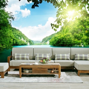 May include: A light brown wooden sectional sofa with beige cushions and plaid throw pillows sits in front of a scenic mural depicting a lake surrounded by lush green trees and mountains. A matching coffee table with a wooden top and a lower shelf sits in front of the sofa.