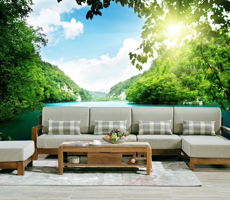 May include: A light brown wooden sectional sofa with beige cushions and plaid throw pillows sits in front of a scenic mural depicting a lake surrounded by lush green trees and mountains. A matching coffee table with a wooden top and a lower shelf sits in front of the sofa.