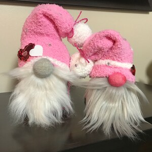 May include: Two pink sock gnomes with white fur beards and pink hearts on their hats. The gnomes are sitting on a brown surface.