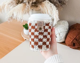 May include: A crochet coffee cup sleeve featuring a checkered pattern in rust orange and cream white. The sleeve has a white plastic lid and a small tan leather tag with the text Cozy Dahlia Designs attached by a brass-colored metal rivet.