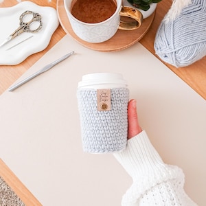 May include: A light blue knitted coffee cup sleeve with a leather tag that reads "Cozy Cabin Designs". The sleeve is wrapped around a white disposable coffee cup. A cup of coffee, scissors, and yarn are also visible.
