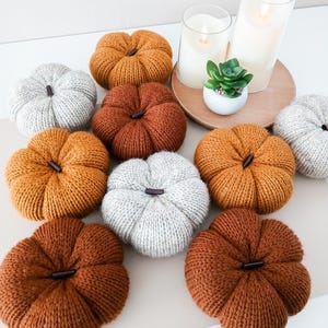 May include: A collection of nine knitted pumpkins in shades of orange, rust, and light gray, each featuring a dark brown stem. Two cylindrical glass candles and a small green succulent in a white pot are arranged on a round wooden tray.