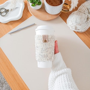 May include: A white paper coffee cup with a knitted cosy. The cosy is white and grey with a small leather tag that reads "Cozy Darlin Designs". The cup is held by a person wearing a white sweater.