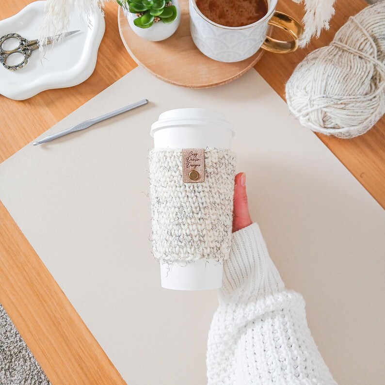 May include: A white paper coffee cup with a knitted cosy. The cosy is white and grey with a small leather tag that reads "Cozy Darlin Designs". The cup is held by a person wearing a white sweater.