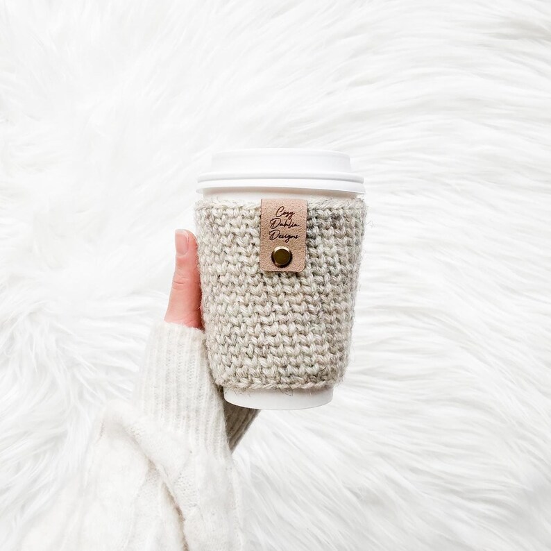 May include: A beige knitted coffee cup sleeve with a leather tag that reads "Cozy Dublin Designs". The sleeve is wrapped around a white disposable coffee cup with a white lid. The sleeve is held by a person.