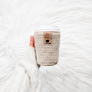 May include: A beige knitted coffee cup sleeve with a white lid. A small brown leather tag with the text "Cozy Dublin Designs" is attached. The sleeve is held by a person wearing a white sweater.