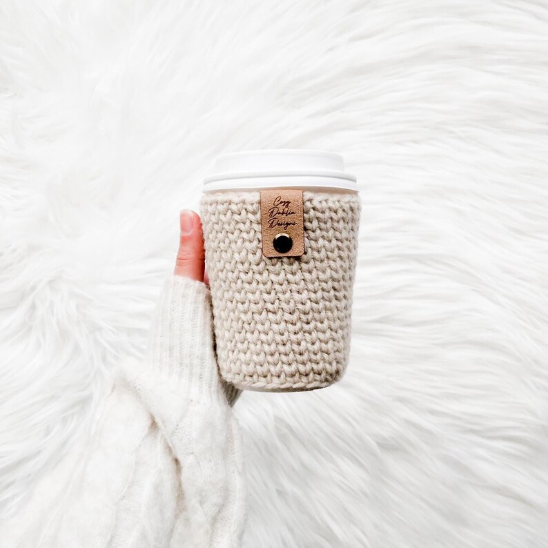 May include: A beige knitted coffee cup sleeve with a white lid. A small brown leather tag with the text "Cozy Dublin Designs" is attached. The sleeve is held by a person wearing a white sweater.