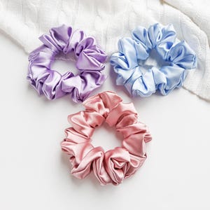 May include: Three satin scrunchies in different colors: lavender, light blue, and pink. The scrunchies are arranged on a white surface.