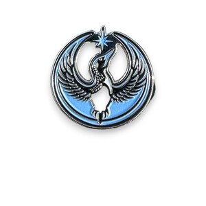 May include: A round, enamel pin featuring a stylized bird with outstretched wings, a star, and a light blue background. The bird is black and white with a red eye, set against a silver-colored metal border.