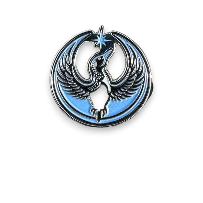 May include: A round, enamel pin featuring a stylized bird with outstretched wings, a star, and a light blue background. The bird is black and white with a red eye, set against a silver-colored metal border.