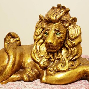 May include: A golden lion statue in a reclining pose. The sculpture has a detailed mane, a regal expression, and green eyes. The lion is resting on a patterned surface, showcasing its decorative nature.