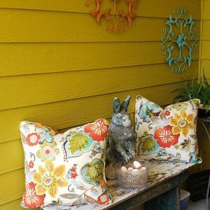 May include: A rustic wooden bench with floral pillows and a gray rabbit statue. The bench is set against a yellow wall adorned with decorative metal wall art. A metal watering can sits on the ground.