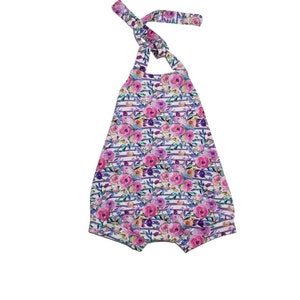 May include: A white baby romper with a floral print featuring pink, purple, and blue flowers. The romper has a halter neck style with a tie at the back.