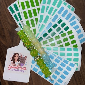 Puede incluir: Imagen de accesorios para pintura de diamantes. Muestra dos bolígrafos con purpurina, cartas de colores y una etiqueta blanca con el texto "Sparkle With Liz And Benny" y una ilustración de una mujer y un perro.