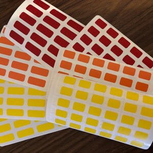Puede incluir: Varias hojas de pegatinas con un patrón rectangular repetido en rojo, naranja y amarillo. Las pegatinas están dispuestas sobre una superficie de madera oscura.