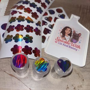 Op de afbeelding: Een verzameling holografische stickers en containers. De stickers hebben bloem-, hart- en cirkelvormen in iriserende kleuren. De containers bevatten vergelijkbare holografische vormen. Er is ook een witte kaart met de tekst "Sparkle With Liz And Benny".