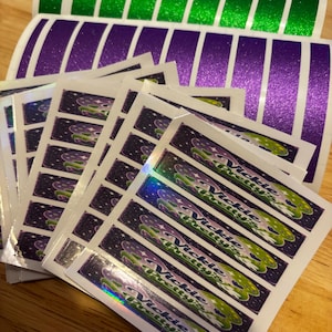 Puede incluir: Una colección de pegatinas rectangulares con un fondo morado oscuro con estrellas blancas y un elemento gráfico verde. Cada pegatina muestra el texto "Vickie Pretty's" en fuente blanca. El conjunto incluye hojas de tiras verdes y moradas sólidas.