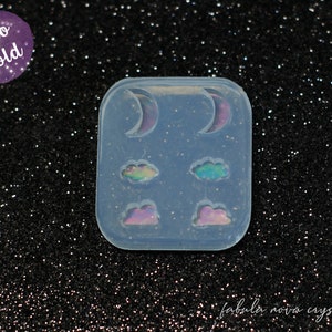 Puede incluir: Un molde de silicona transparente con cuatro cavidades para hacer pendientes de resina. El molde presenta dos formas de luna creciente y dos formas de nube. El molde está etiquetado como "Holo Mold".