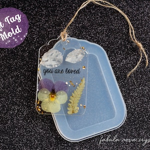 May include: A clear silicone mold shaped like a gift tag with a hole at the top for a string. The mold is filled with resin and includes dried flowers, a fern, gold leaf, and a clear quartz crystal. The resin is clear and the flowers are purple, yellow, and green. The text "you are loved" is visible in the resin. The mold is on a black surface.