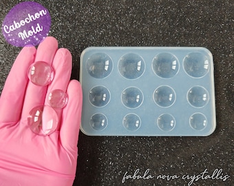 Könnte beinhalten: Eine transparente Cabochon-Form mit mehreren kreisförmigen Hohlräumen, zusammen mit mehreren klaren, gewölbten Cabochons. Der Text "Cabochon Mold" befindet sich in einem lila Kreis. Der Text "fabula nova crystallis" steht unten.
