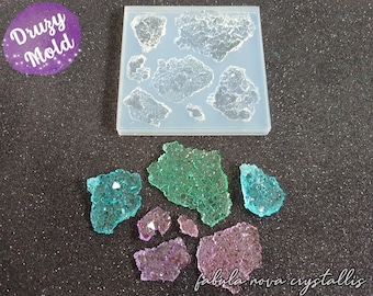May include: A clear silicone mold with various crystal shapes, alongside several finished resin crystals in teal, green, and purple. The image includes the text "Druzy Mold" and "fabula nova crystallis".