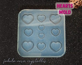 May include: A clear, flexible silicone mold with nine heart-shaped cavities. The mold is labeled "HEARTS MOLD" and "fabula nova crystallis". The hearts vary in size, perfect for crafting resin jewelry, charms, or decorative elements. The mold is set against a dark, glittery background.