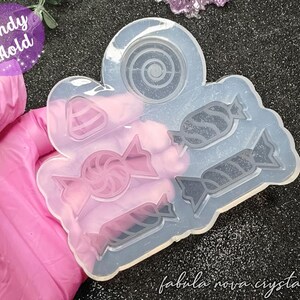 Puede incluir: Un molde de silicona transparente para caramelos con varias formas de caramelos, incluyendo una piruleta y caramelos envueltos. El molde está sostenido por una mano enguantada. El texto "Candy Mold" es visible en un círculo púrpura.