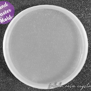 May include: A clear, round coaster mold with a slightly textured interior. The mold has a raised edge and is labeled "Round Coaster Mold" in a purple circle. The words "fabula nova crystallis" are written in cursive at the bottom.